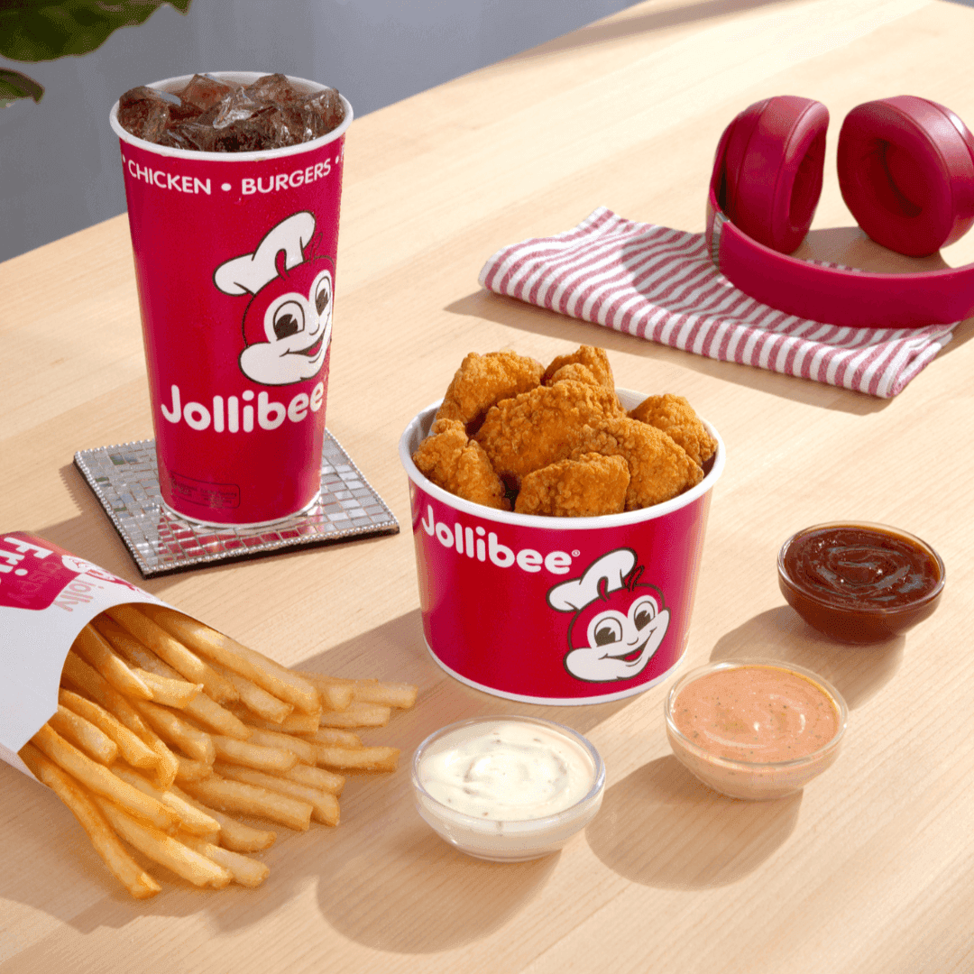 A red bucket of chicken nuggets with the Jollibee logo on it, with a drink in a red cup, a side of fries, and three different dips on a table.