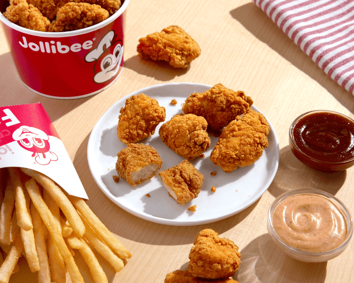 Chicken nuggets pieces on a white plate and some inside the red bucket with a side of fries and two dipping sauces on the table.