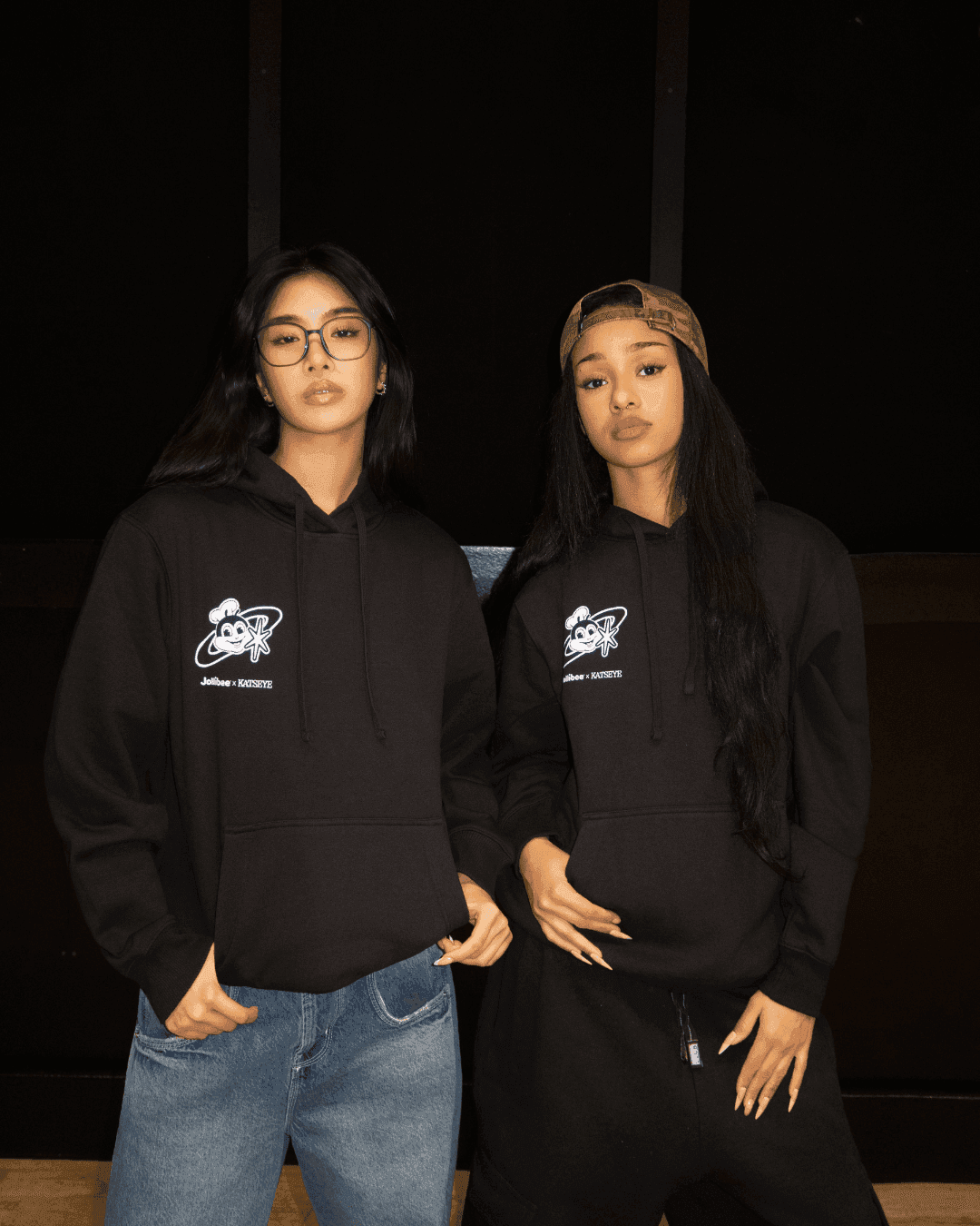 Two members of the KATSEYE girl group wearing black hoodies with logos, standing against a dark background.