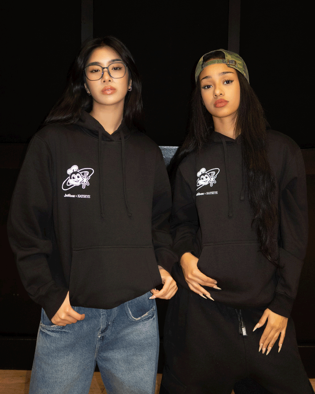 Two girls wearing a black hoodie with the Jollibee x KATSEYE shared dream logo on the chest in a dark background