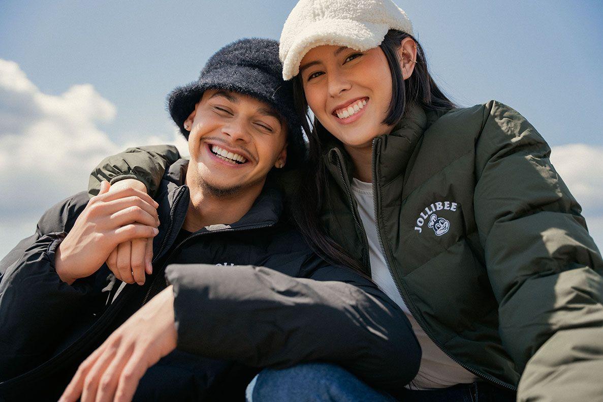 Two people wearing Jollibee jackets and beanies, smiling outdoors with a clear sky.
