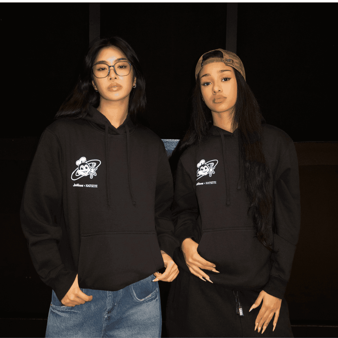 Two members of the KATSEYE girl group wearing black hoodies with logos, standing against a dark background.